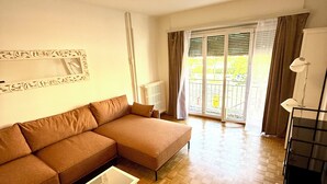 Un salón con un sofá seccional, una mesita blanca, un radiador y vistas al exterior a través de un ventanal.