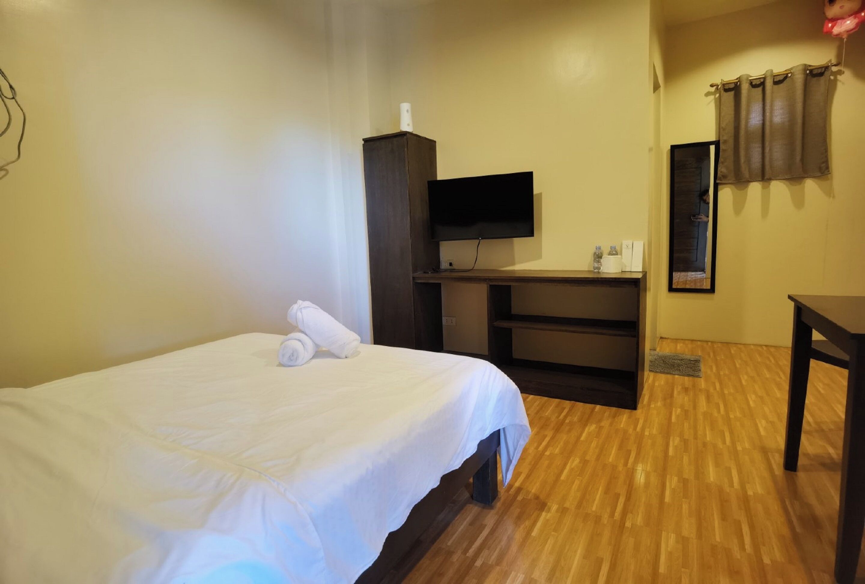 A hotel room with a bed, a TV mounted on a wooden cabinet, a small desk, and a mirror on the wall.