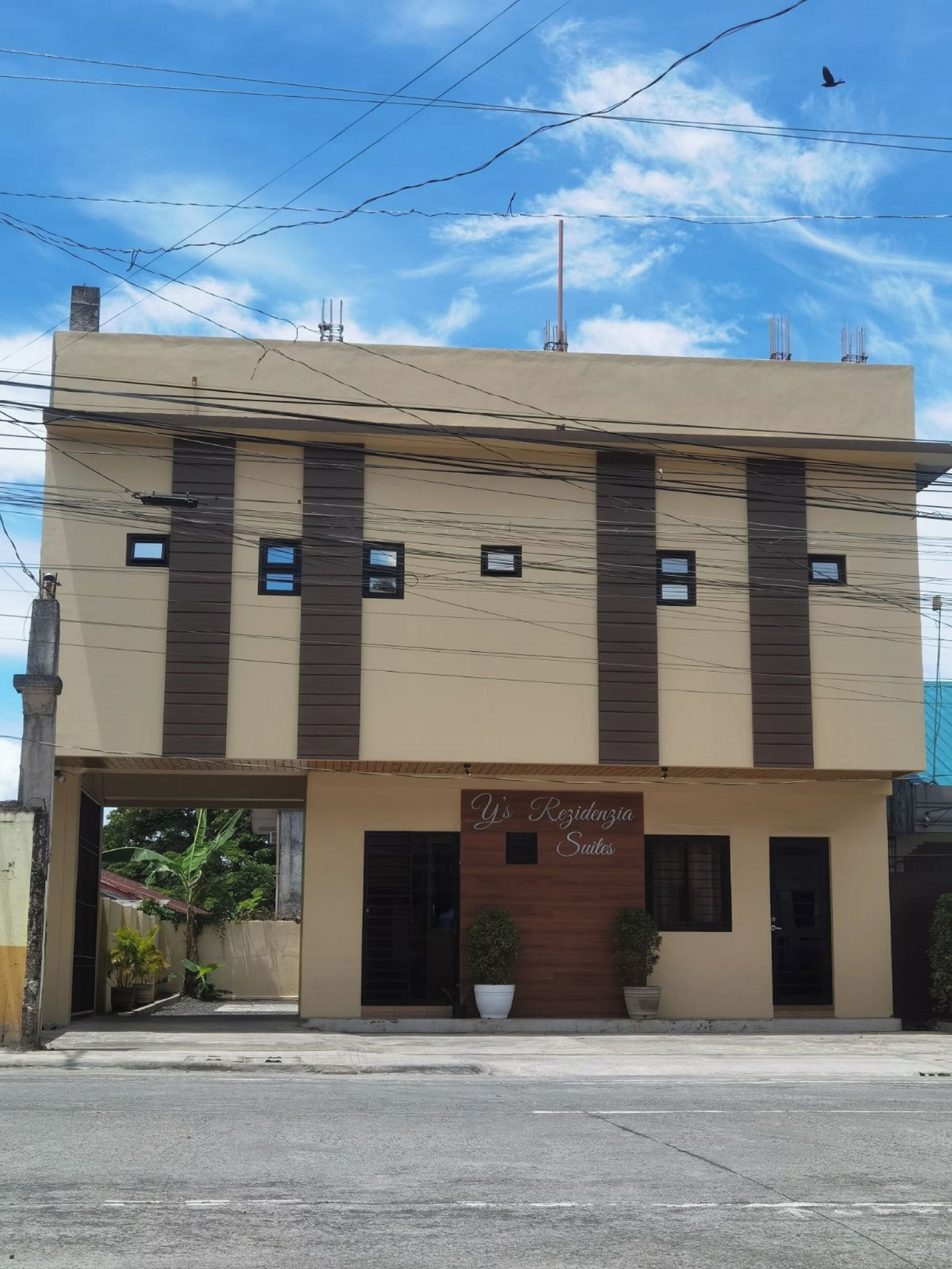 A two-story building with a sign reading "Y's Residencia Suites". There are multiple windows on each floor, and the building has a flat roof.
