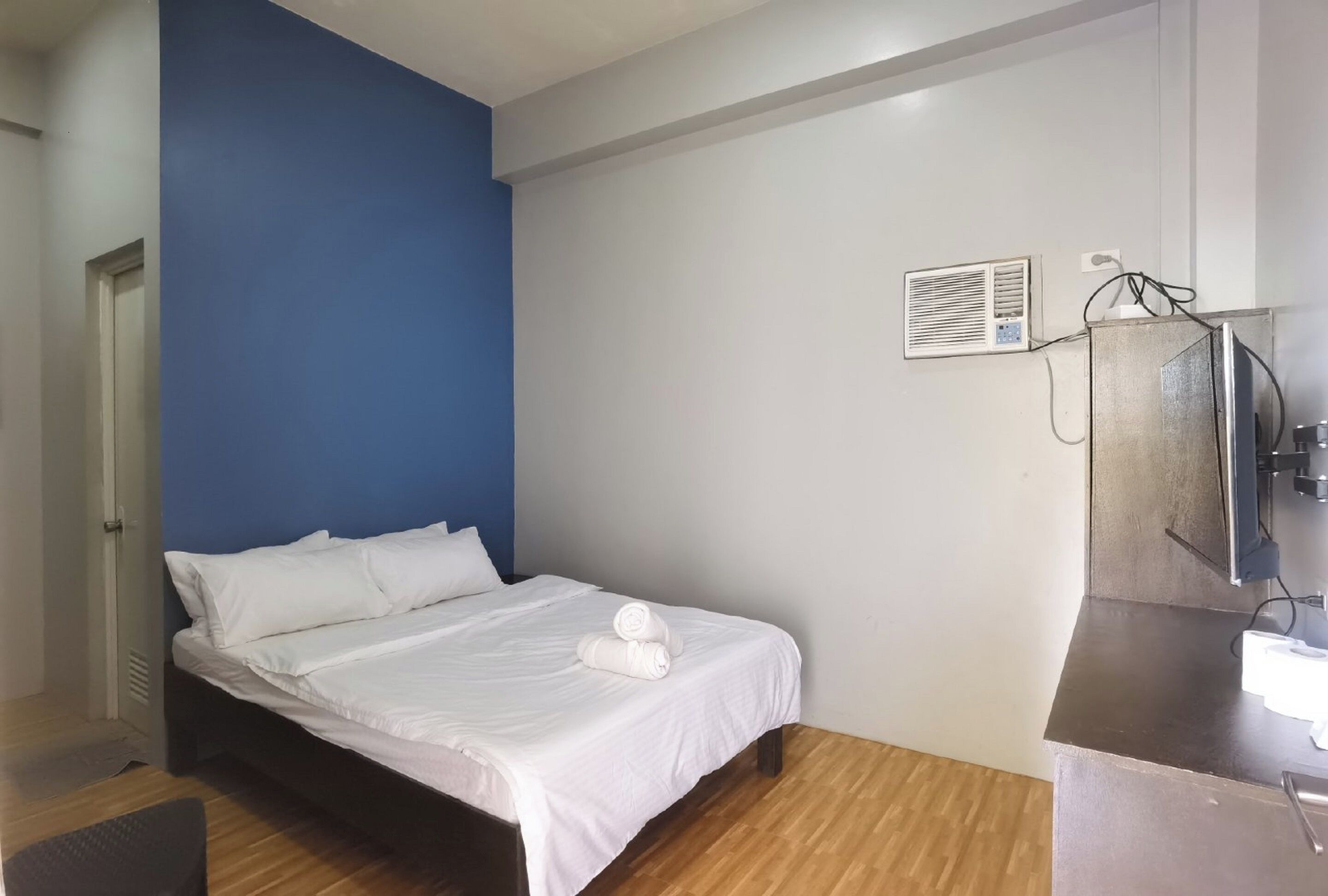 A bedroom with a bed, a blue accent wall, a small desk with a mirror, and an air conditioning unit.