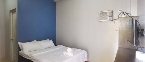A bedroom with a bed, a blue accent wall, a small desk with a mirror, and an air conditioning unit.