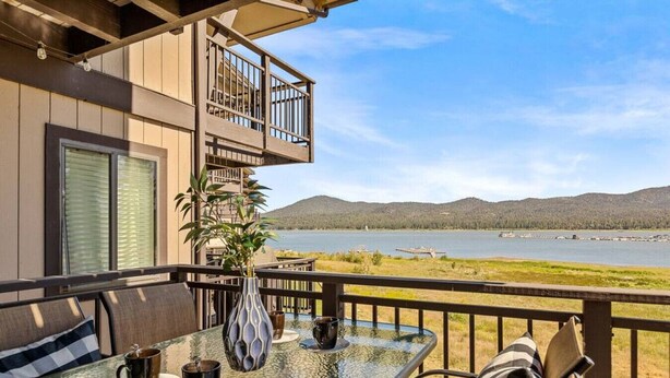 Ein Balkon mit Tisch für zwei Personen, einem Blumentopf und Blick auf einen See und Berge.
