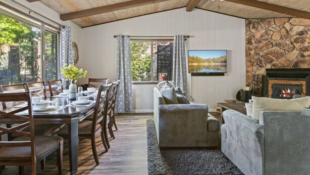 A dining area with a wooden table and chairs, a living room with a fireplace, and a stone wall. - Charming, cozy and rustic cabin! (Big Bear)