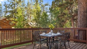 A wooden deck with a table and chairs, surrounded by trees and a house in the background. - Charming, cozy and rustic cabin! (Big Bear)
