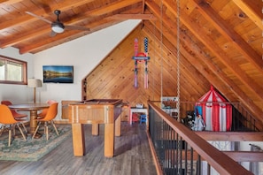 A loft-style room with a wooden ceiling, a pool table, orange chairs, and a red and white striped tent.