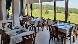 Un restaurant avec des tables dressées pour le service, de grandes fenêtres donnant sur un paysage verdoyant.