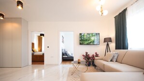 A modern living room with a beige sofa, a coffee table, and a painting of a forest. There is a door leading to another room and a floor lamp.