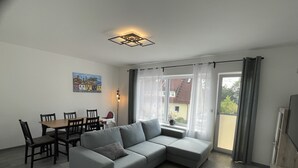 A modern living room with a grey sectional sofa, a dining table with chairs, a small round table, and a large window with curtains. - Quiet accommodation with best location for trips to Nuremberg and surroundings  (Feucht)