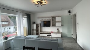 A modern living room with a grey sofa, a dining table with chairs, and a kitchen area with cabinets and a TV. - Quiet accommodation with best location for trips to Nuremberg and surroundings  (Feucht)