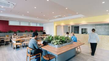 A modern cafe with wooden tables and chairs, a central plant display, and a red accent wall.