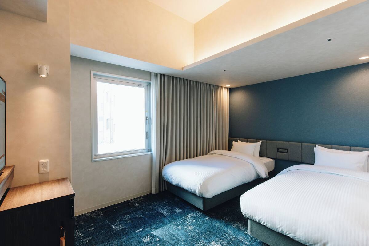 A hotel room with two beds, a window with curtains, a desk, and a blue carpet.