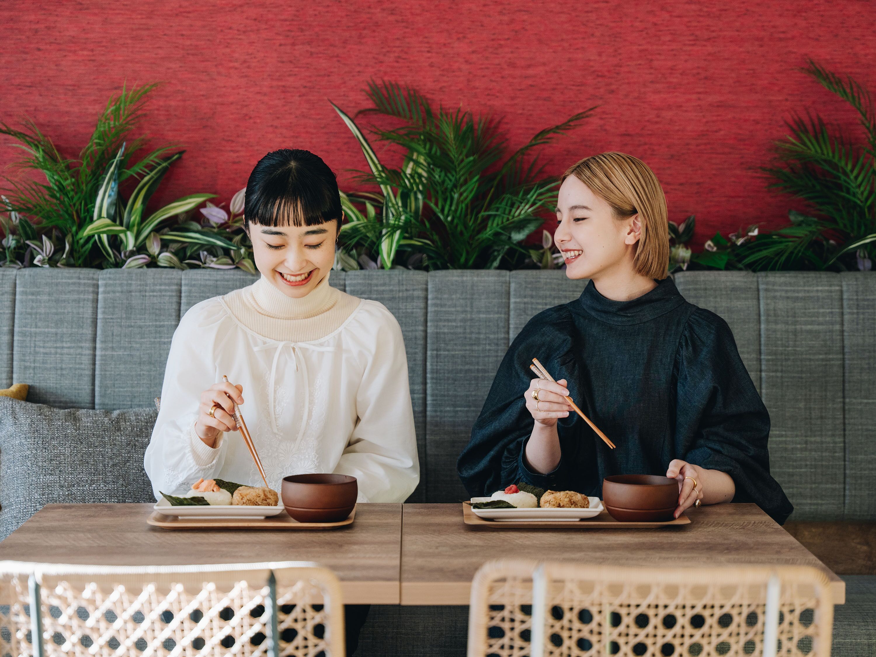 Two people sitting at a table with bowls of food, a grey sofa, and green plants in the background.