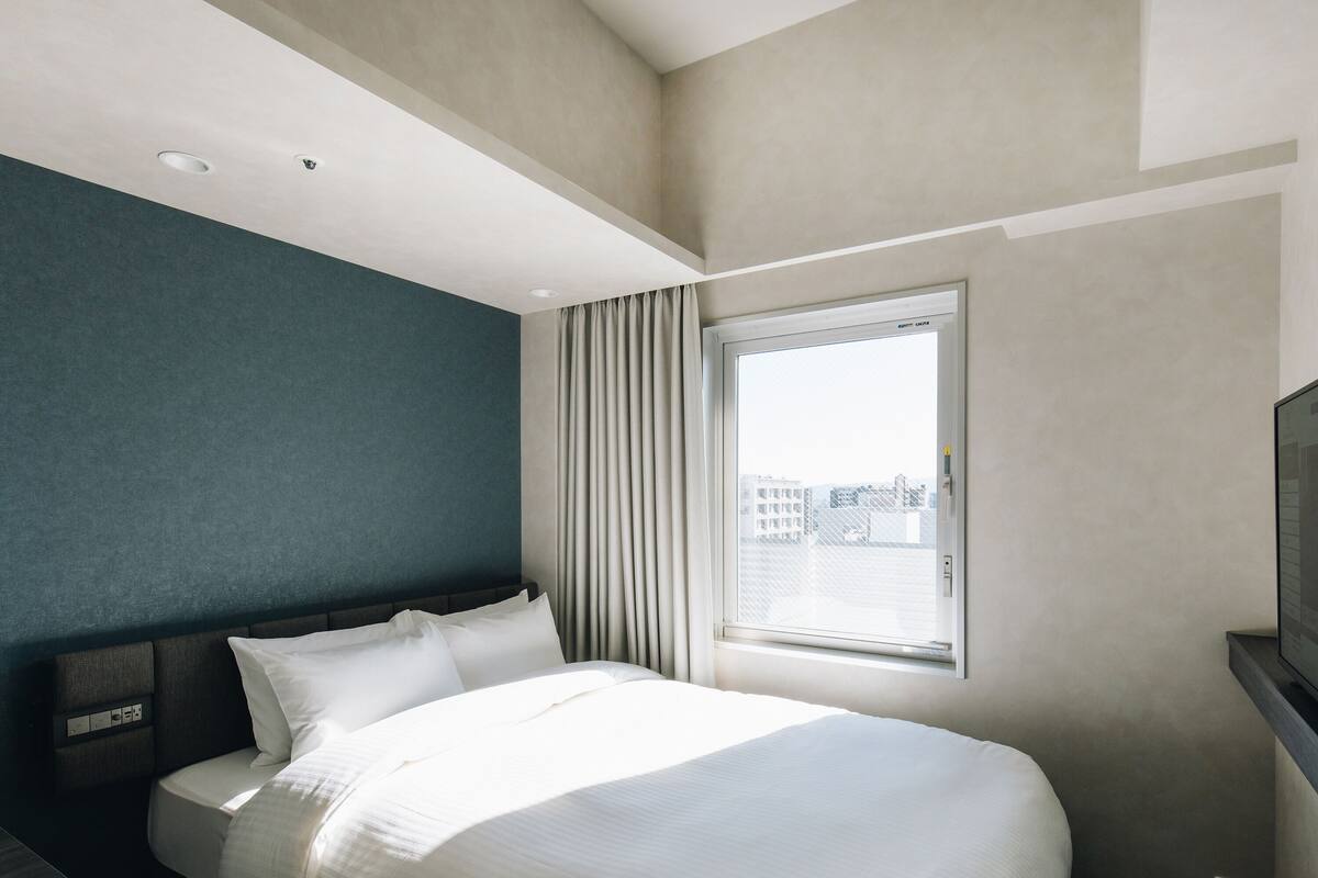 A hotel room with a bed, a window with curtains, a television, and a wall with a textured design.