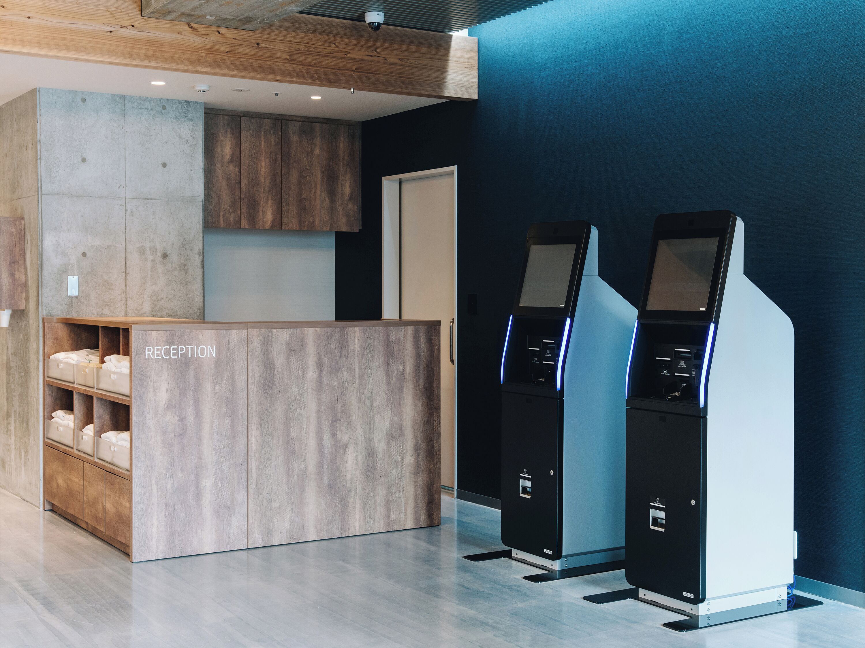 A reception area with a wooden reception desk, a blue wall, and two self-service kiosks.