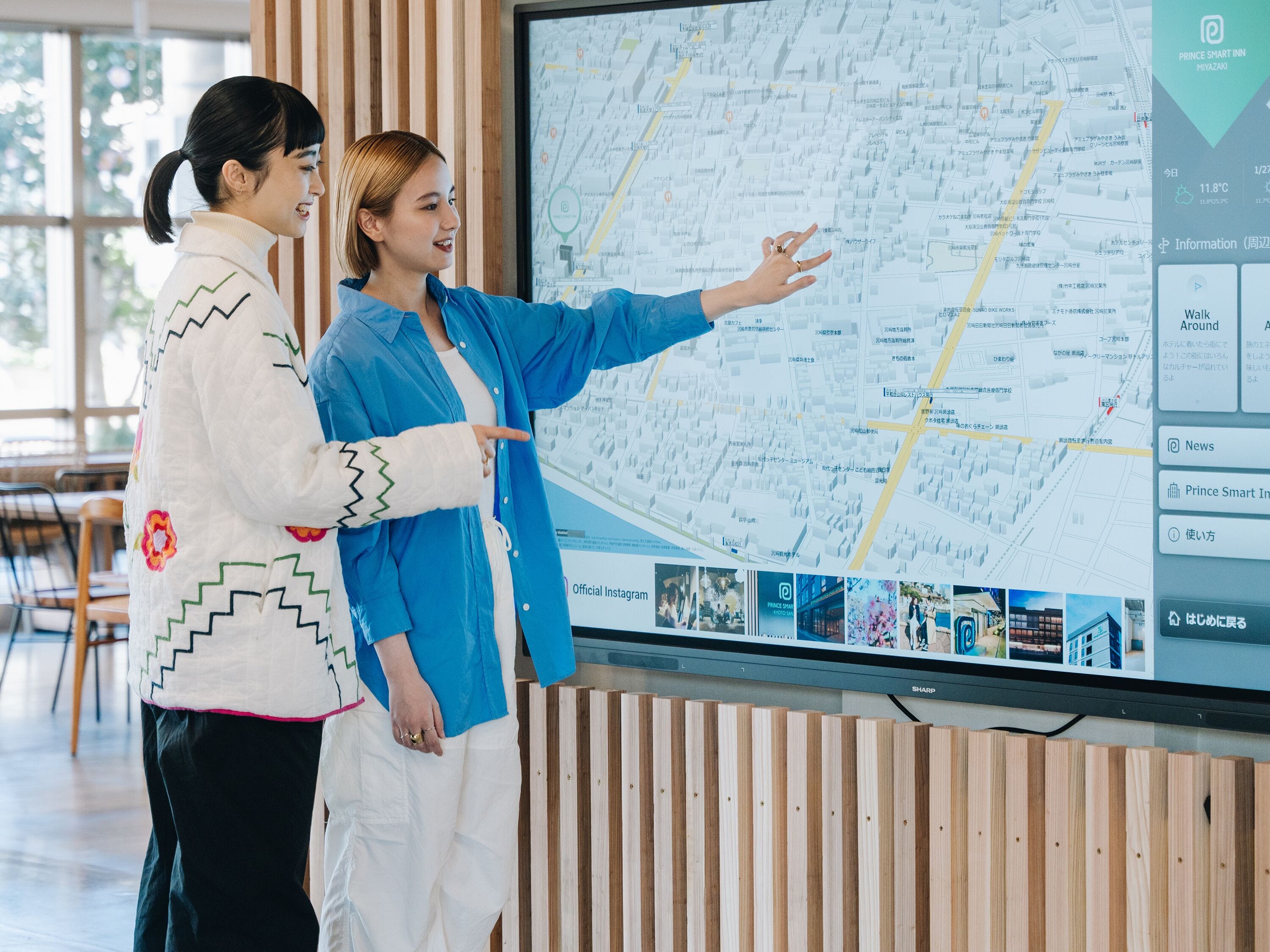 Two individuals examining a large map on a screen, with a wooden panel and a window in the background.