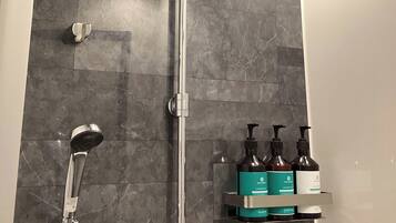 A shower area with a glass door, a handheld showerhead, and a shelf with three bottles of shampoo and conditioner.