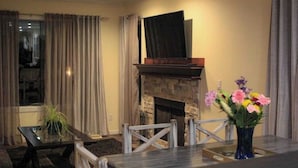 Una sala de estar con chimenea, un televisor de pantalla plana montado encima, una mesa de comedor con sillas y un florero con flores.