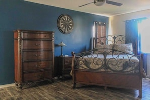 A bedroom with a wooden dresser, a bed with ornate metal headboard, a wall clock, and a ceiling fan.