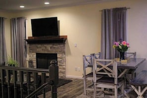 A living room with a stone fireplace, a flat-screen TV mounted above it, and a dining area with wooden chairs and a table.