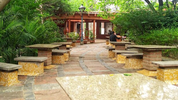 A garden pathway with stone benches and tables, surrounded by lush greenery and a red-roofed structure. - Roomshala 087 Green High (New Delhi)