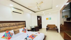 A hotel room with a bed, a TV, a ceiling fan, an air conditioning unit, a desk, a chair, and a door. - Roomshala 087 Green High (New Delhi)