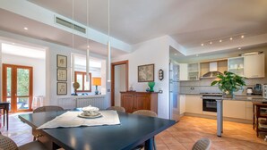 Un vaste espace de salle à manger avec une grande table et des chaises, une cuisine dotée de meubles blancs et une vue sur l’extérieur.