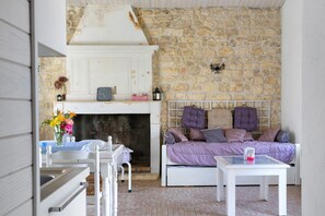 A room with a stone wall, a white bed with purple bedding, a small table, and a white chair.
