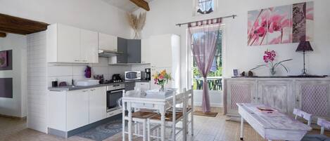 A kitchen with white cabinets, a dining table, and chairs. There is a window with curtains, a flower vase, and a lamp on a cabinet.