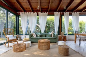 Una habitación con sofá verde, muebles de mimbre y amplias ventanas con cortinas blancas.