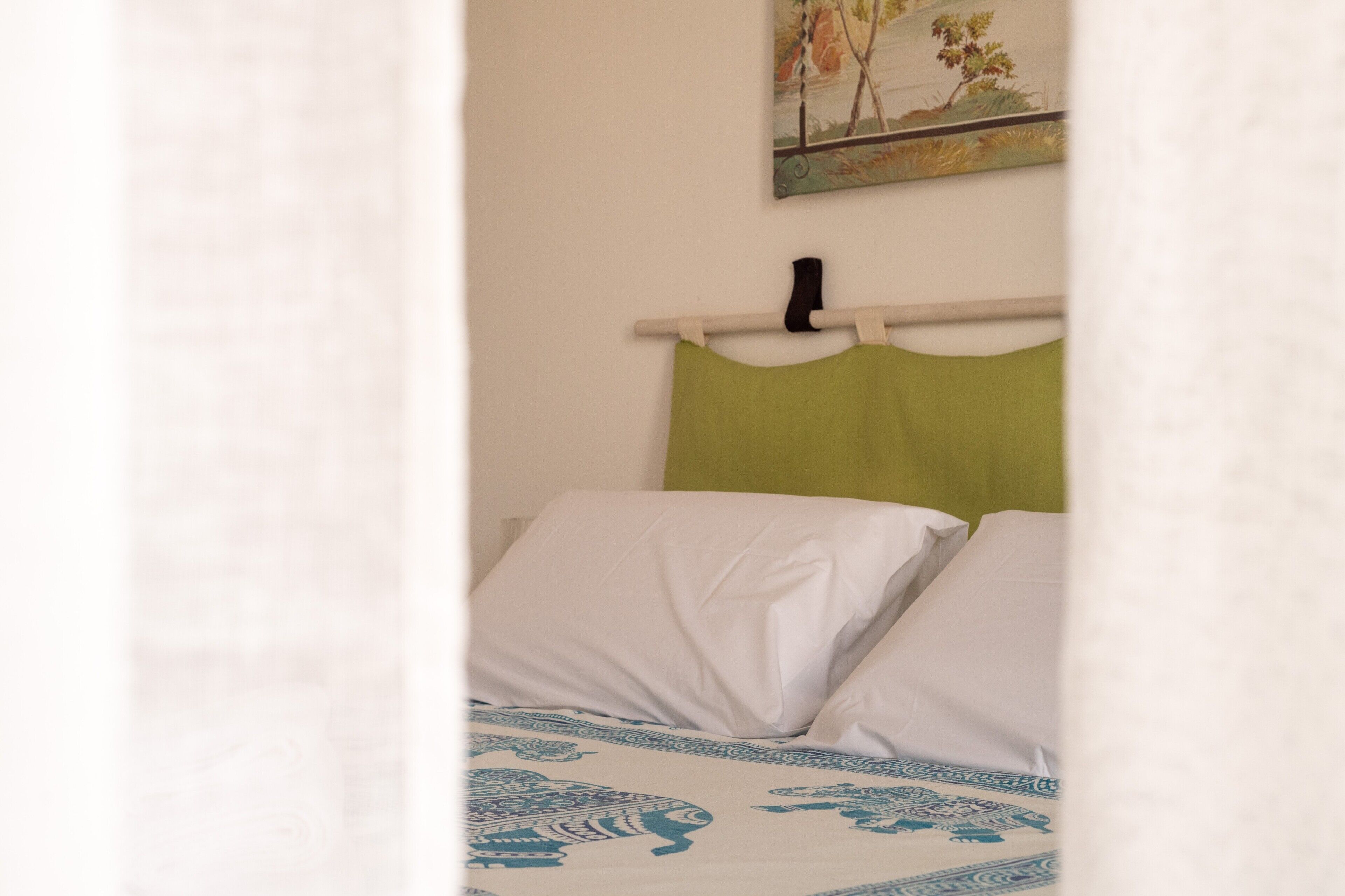 Een bed met wit en groen beddengoed, een schilderij aan de muur en een gedeeltelijk zichtbaar wit gordijn.