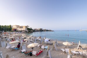 Une plage avec des transats et des parasols, un ciel dégagé et des bateaux sur l’eau.