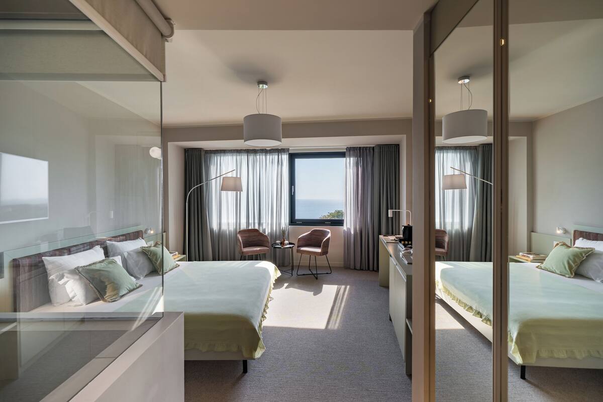 A hotel room with two beds, a desk, a chair, and a view of the outdoors.