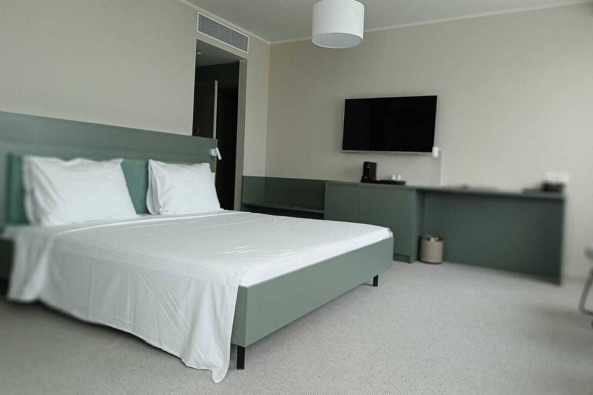 A modern hotel room with a single bed, a flat-screen TV mounted on the wall, and a small kitchenette area.