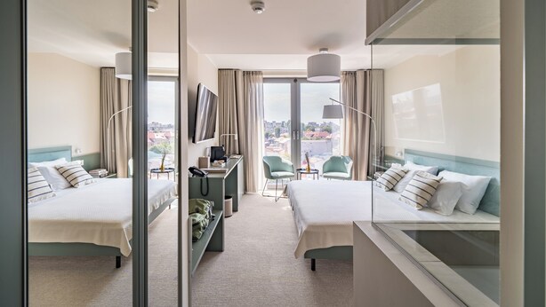 A hotel room with two beds, a desk, a chair, a TV, and a view of the city. - MILLENNIUM (Constanta)