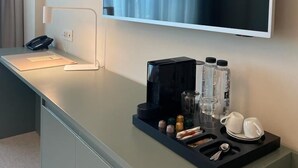 A hotel room with a flat-screen TV mounted on the wall, a desk with a phone, a lamp, and a tray table with water bottles, glasses, and a coffee pot. - MILLENNIUM (Constanta)
