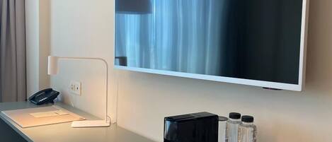 A hotel room with a flat-screen TV mounted on the wall, a desk with a phone, a lamp, and a tray table with water bottles, glasses, and a coffee pot.