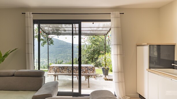 A modern living room with a sofa, a dining table, and a view of a mountain landscape through large glass doors. - Apartment "Gîte les Monteils" with Mountain View, Private Terrace & A/C (Vesseaux)
