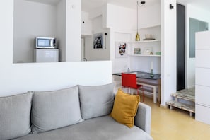 Uma sala de estar moderna com um sofá cinzento, um travesseiro amarelo mostarda, uma cadeira vermelha e uma área de cozinha com micro-ondas e uma mesa de jantar.