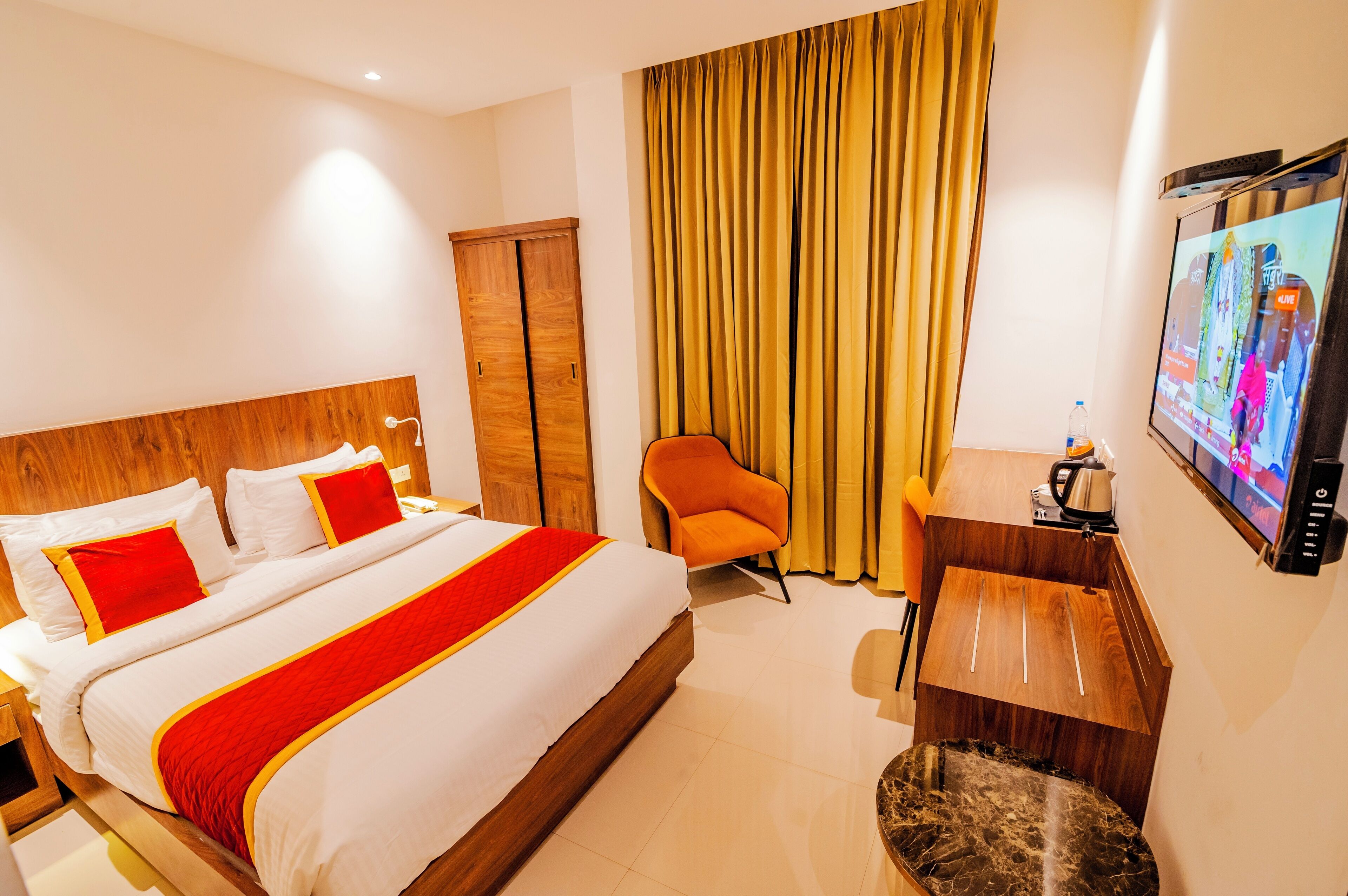 Habitación de hotel con una cama grande, una silla naranja, un escritorio con cafetera y un televisor en la pared.