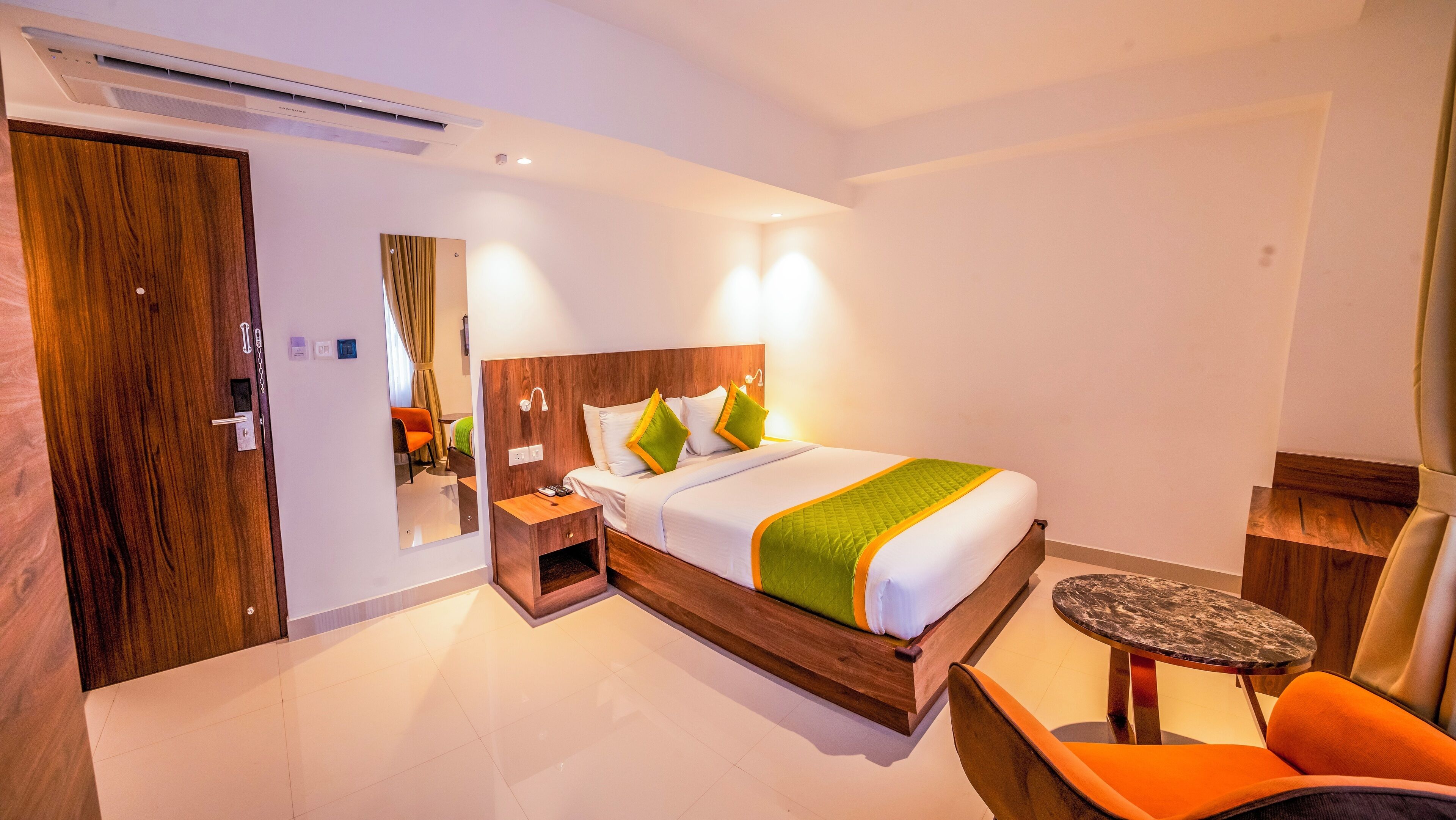 A hotel room with a large bed, a wooden nightstand, a chair, a small table, and a mirror.