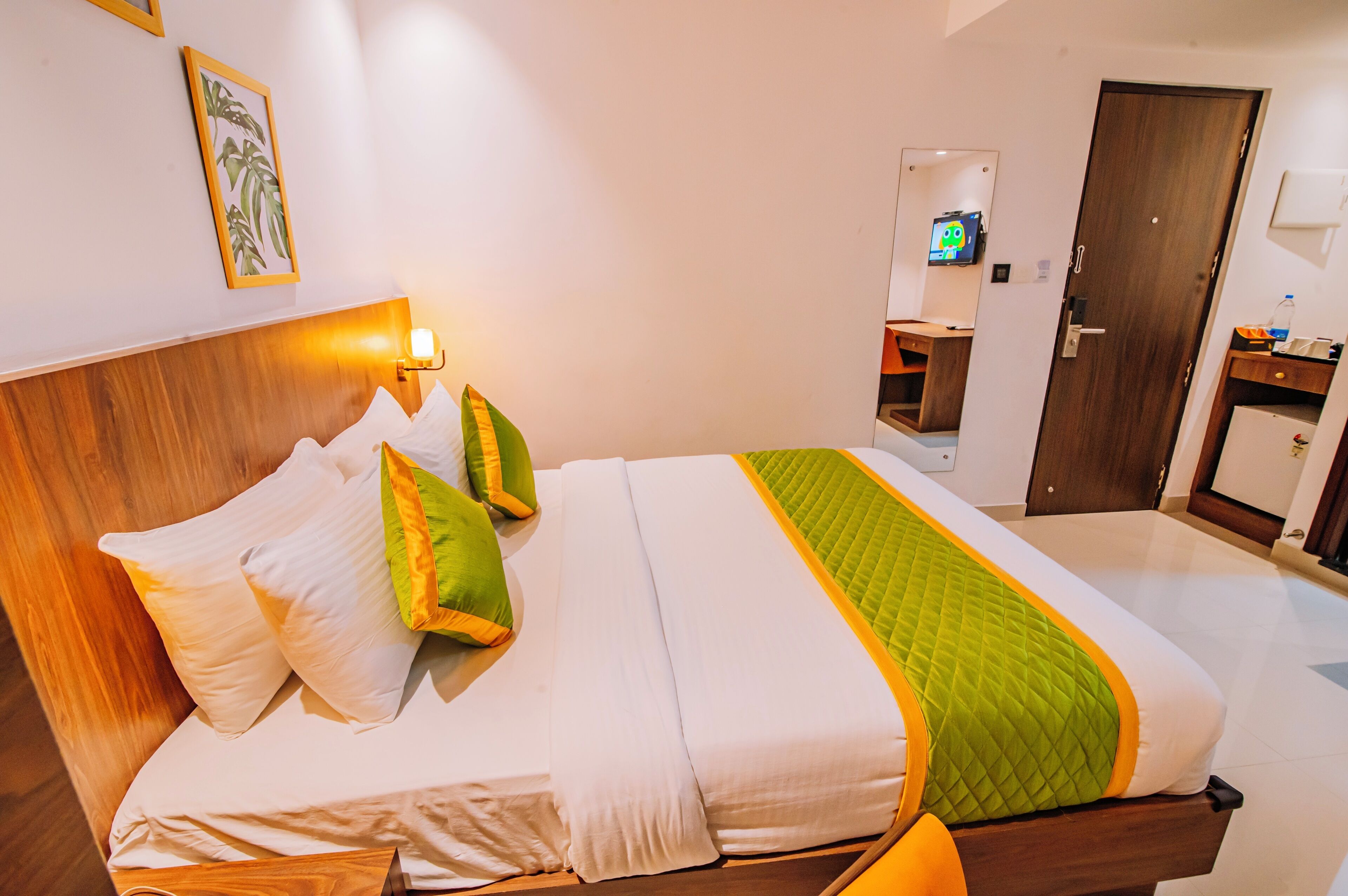 A hotel room with a wooden headboard, a bed with white and green bedding, a bedside table with a lamp, and a door with a mirror and a television.