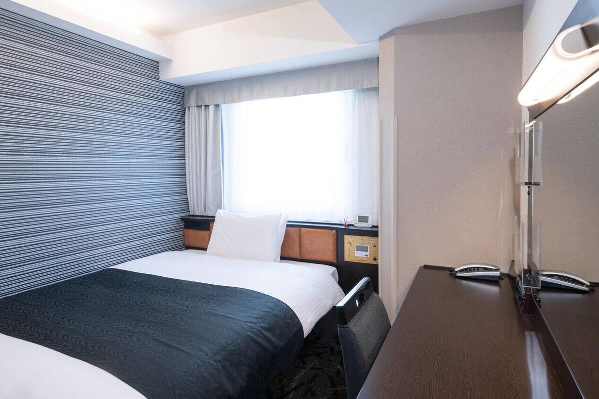 A hotel room with a bed, a desk, a chair, a window with curtains, and a wall with a striped pattern.