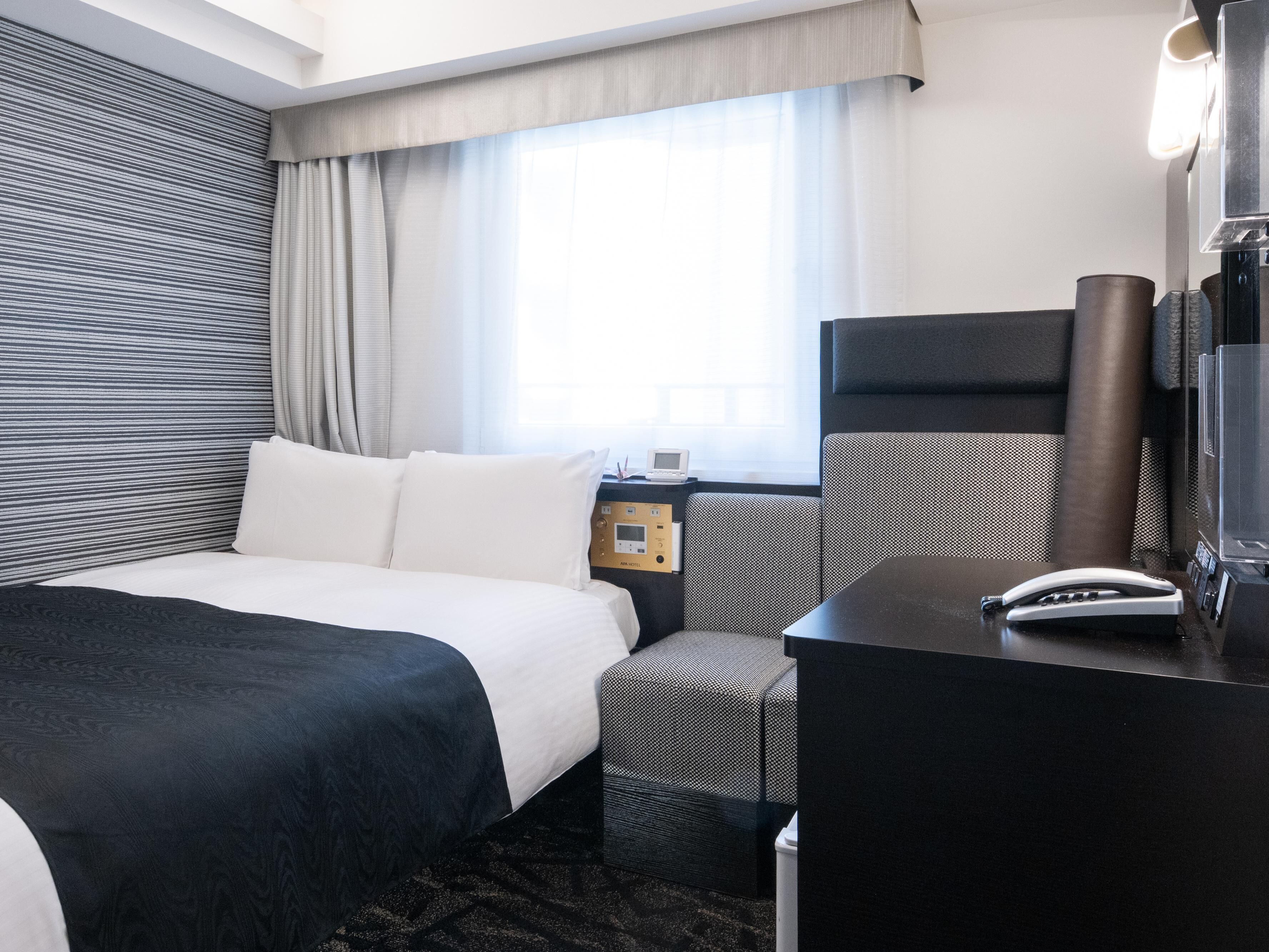 Quarto de hotel com cama, escrivaninha, cadeira, mesa de cabeceira e janela com cortinas.