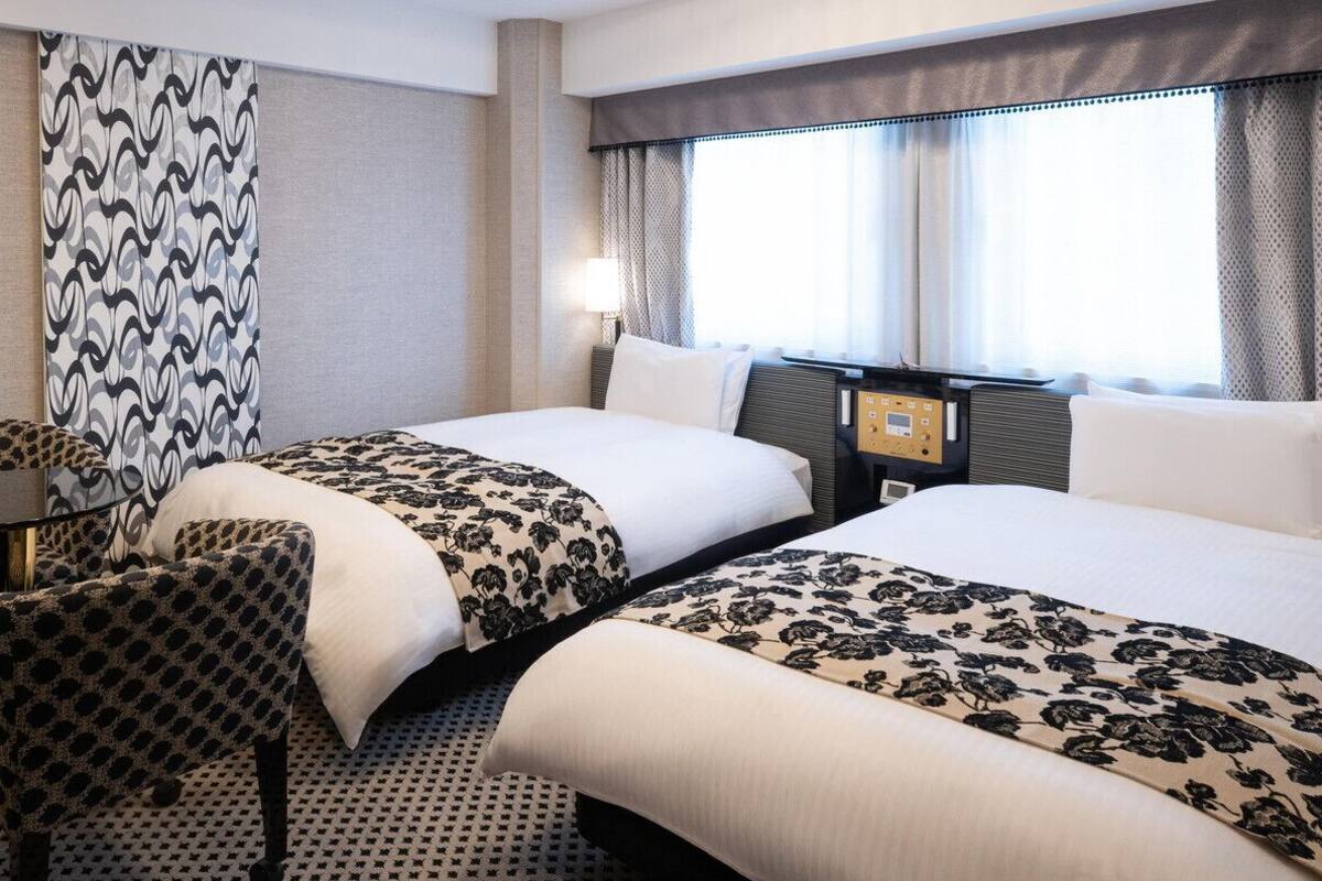 A hotel room with two beds, a chair, a bedside table, and a window with curtains.