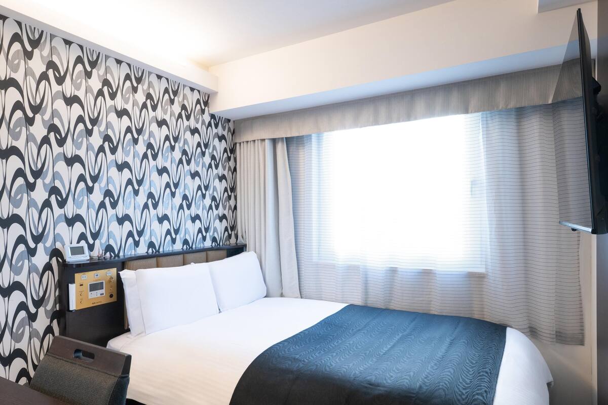 A hotel room with a large bed, a desk with a control panel, a TV, and a window with curtains.