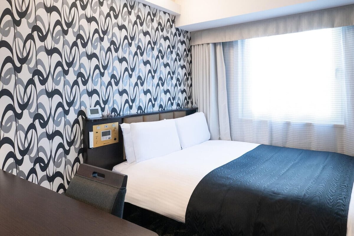 A hotel room with a bed, a bedside table, a control panel, and a window with curtains.