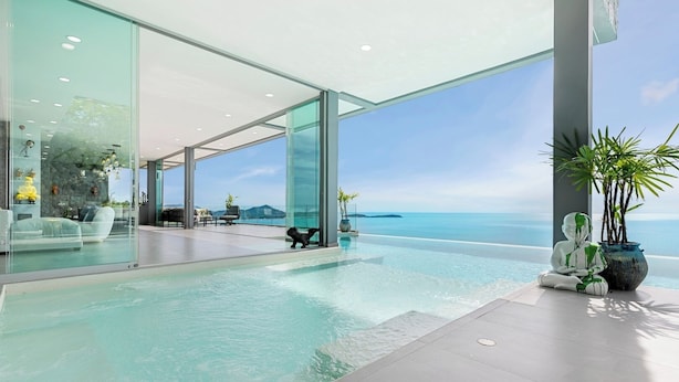 Une piscine à débordement moderne offrant une vue dégagée sur l’océan, un salon vitré et une plante en pot.
