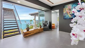 Un intérieur moderne avec un escalier menant à un balcon donnant sur la mer, un coin salon agrémenté de plantes et un grand tableau abstrait.