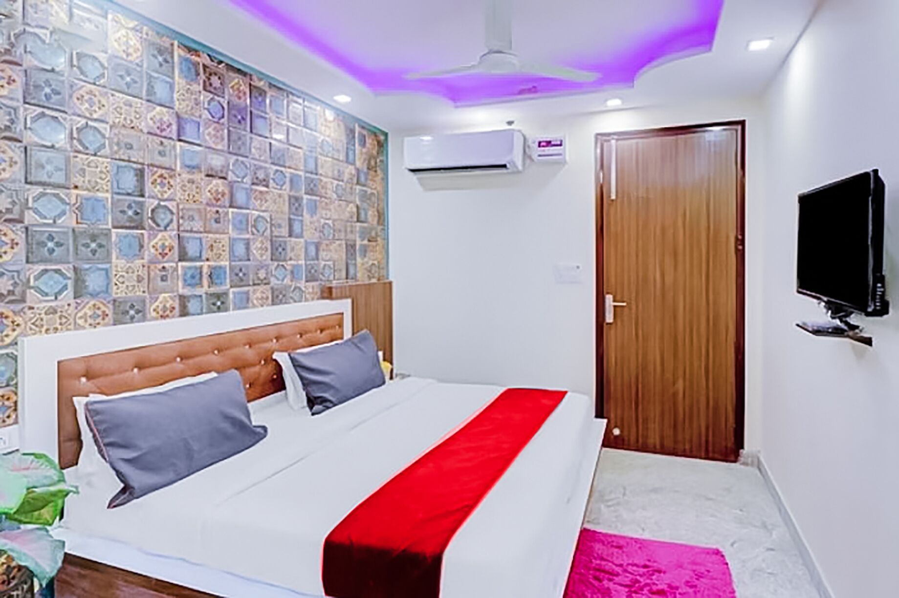 A bedroom with a bed, a wall-mounted air conditioner, a wooden door, and a television on the wall.