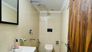 A small, narrow bathroom with a white sink, a toilet, and a shower. There is a wooden door on the right side. - Roomshala 089 Green Palace (New Delhi)